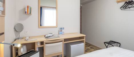 Une chambre d’hôtel comprenant un lit, un bureau avec un miroir, une horloge, un climatiseur, un petit réfrigérateur et un porte-manteau.