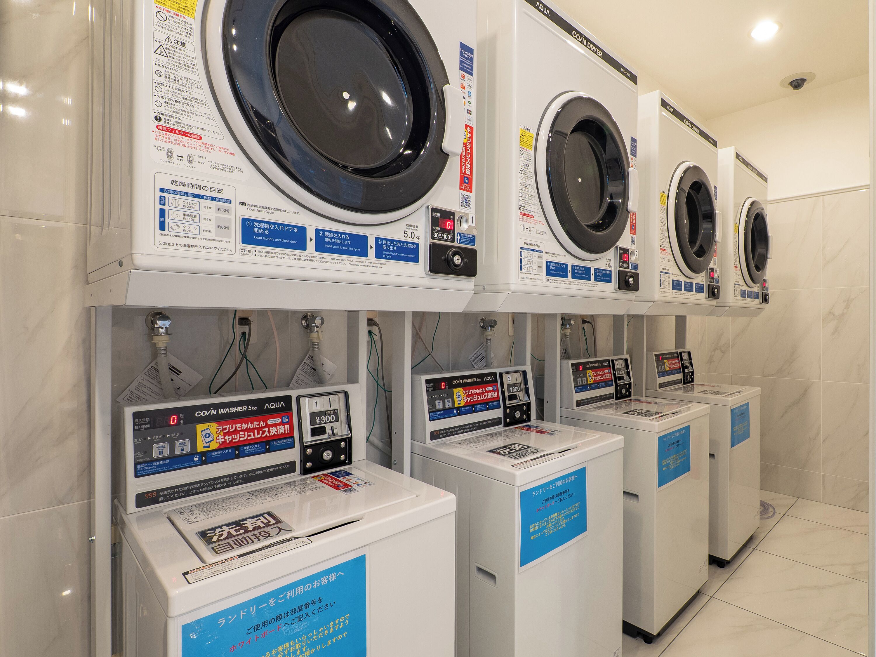 A row of front-loading washing machines and coin-operated dryers in a commercial setting.