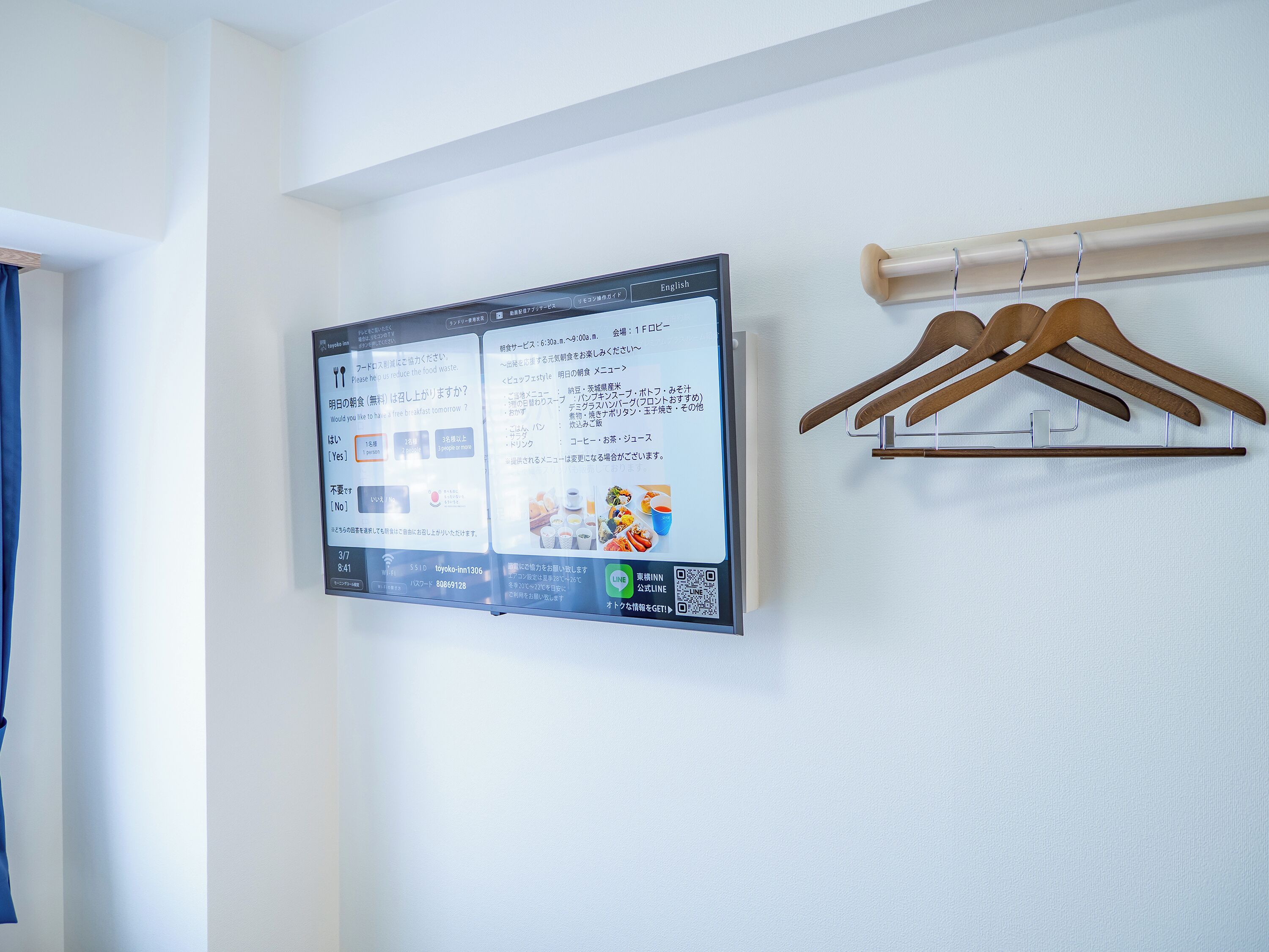 Un panel digital de menú fijado a la pared con texto e imágenes, un perchero de madera con ganchos, y una cortina azul parcialmente visible.