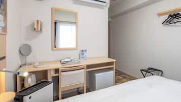 A hotel room with a bed, a desk with a mirror, a clock, an air conditioner, a small fridge, and a coat rack.