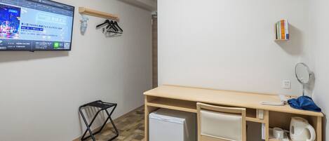 A hotel room with a bed, a desk, a small refrigerator, a wall-mounted TV, an air conditioner, a bookshelf, and a waste bin.