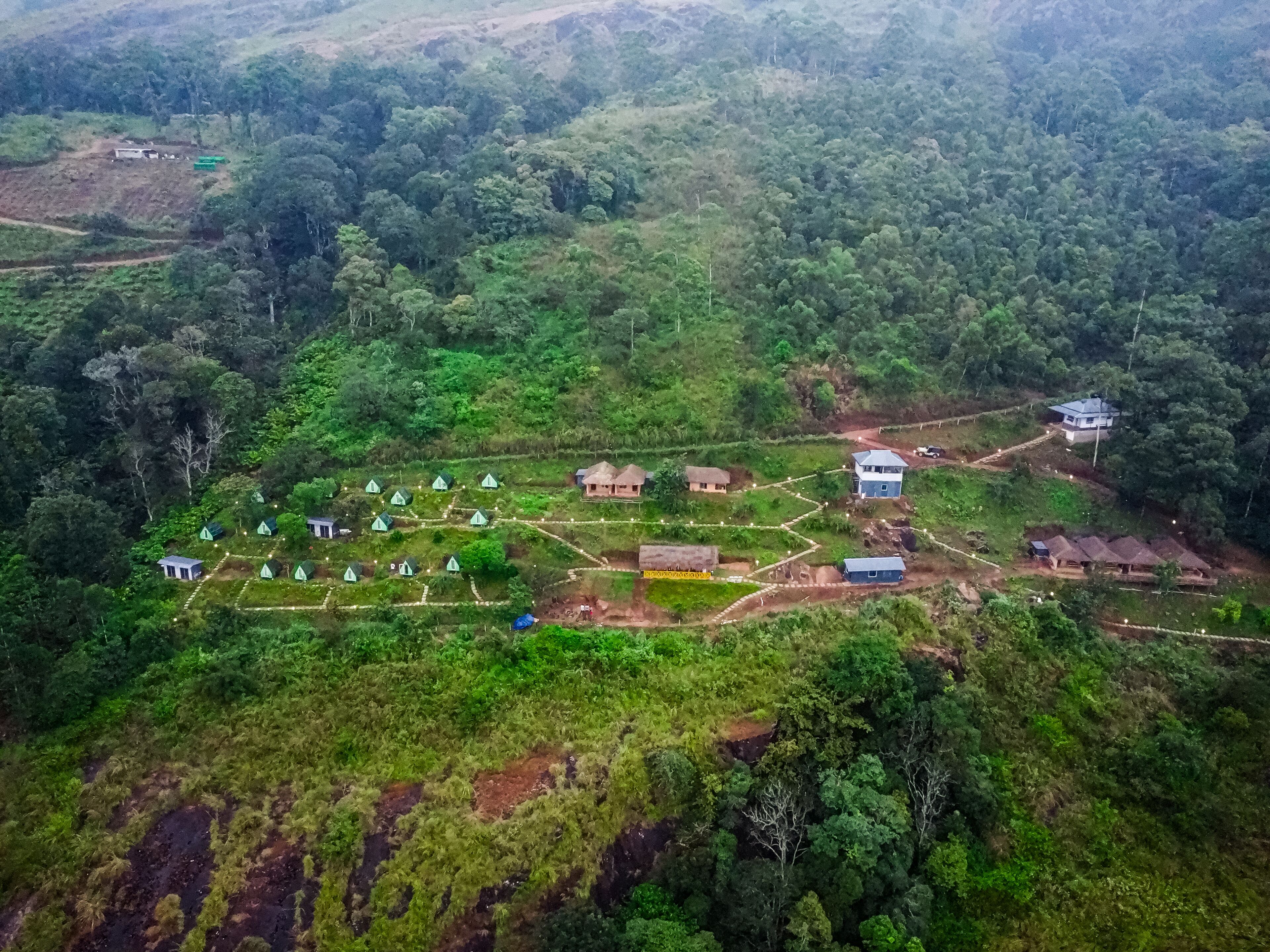 An aerial view of a rural area with several small, traditional-style huts and a few larger buildings, surrounded by dense forest.