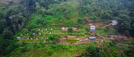 An aerial view of a rural area with several small, traditional-style huts and a few larger buildings, surrounded by dense forest.
