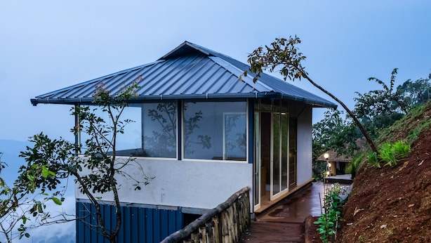 Un sendero de madera lleva a una casa moderna con techo metálico, rodeada de vegetación y con vistas a un paisaje montañoso.