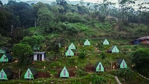Una ladera con varias casitas pequeñas de techo verde, cada una con una ventana y una puerta. Hay senderos que conectan las casitas, y algunas tienen pequeñas luces a lo largo de los caminos.