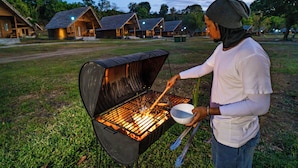 A person cooking on an open grill outdoors with a grassy area and cabins in the background. - Sofanda Resort (Nangamiro)