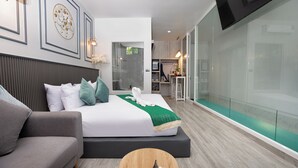 Ein modernes Hotelzimmer mit einem großen Bett, einer Sitzecke, einem Flachbildfernseher und einer Glastrennwand, die den Raum von der Küchenzeile abteilt.