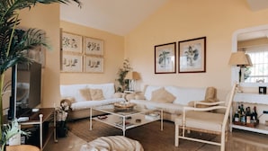 Ein Wohnzimmer mit einem beigefarbenen Sofa, einem Glastisch und einem weißen Sessel. An der Wand befinden sich gerahmte Pflanzenbilder und links eine Topfpflanze.