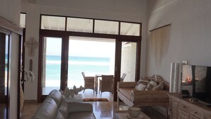 A spacious living room with a large window overlooking the beach, a leather sofa, a wooden coffee table, and a TV on a wooden cabinet.