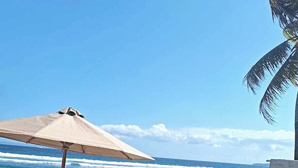 A pool area with lounge chairs and an umbrella, overlooking the ocean with waves crashing onto a stone jetty.