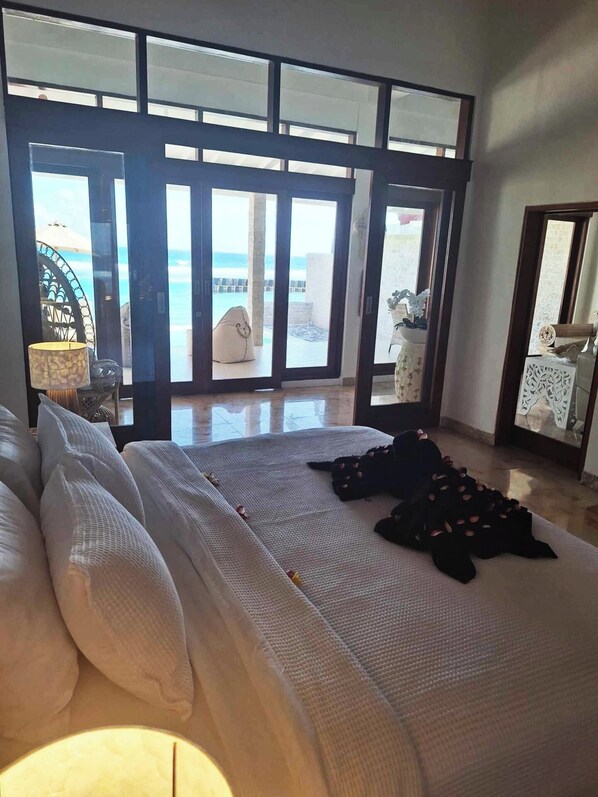 Une chambre avec un grand lit, une table de chevet et une vue sur la plage, visible à travers des portes vitrées.