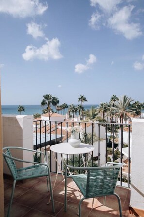 Ein Balkon mit zwei grünen Korbstühlen und einem kleinen runden Tisch, mit Blick auf einen Strand mit Palmen und einem klaren Himmel.