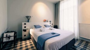 A hotel room with a checkered floor, a bed with white bedding and a blue throw, a bedside table with a lamp, and a window with sheer curtains. - Charming Studio Retreat for Two in Central Ghent (Gent)