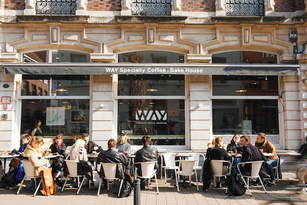 An outdoor cafe with tables and chairs, a sign for 'WAY Specialty Coffee - Bake House,' and a building facade with decorative elements.