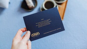 A hand holding a dark blue business card with gold text and a logo, placed on a blue surface with a tray of coffee cups in the background. - Charming Studio Retreat for Two in Central Ghent (Gent)