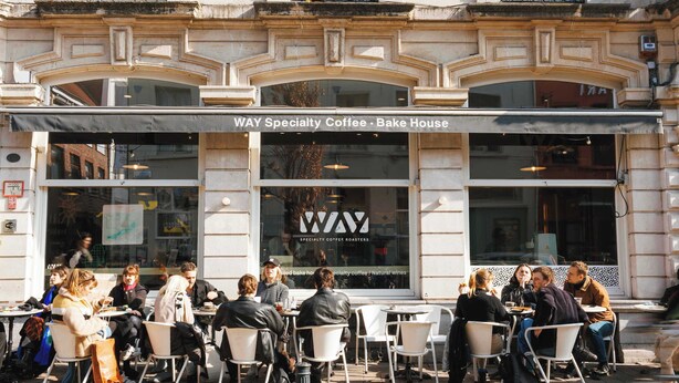 Ein Straßencafé mit Tischen und Stühlen, einem Schild mit der Aufschrift 'WAY Specialty Coffee - Bake House' sowie einer Fassade mit dekorativen Elementen.