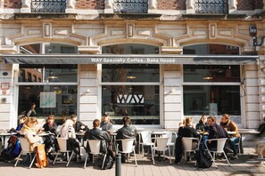 An outdoor cafe with tables and chairs, a sign reading "WAY Specialty Coffee - Bake House," and a building facade with decorative elements. - Ghent Getaway: Cozy Retreat for Two (Gent)