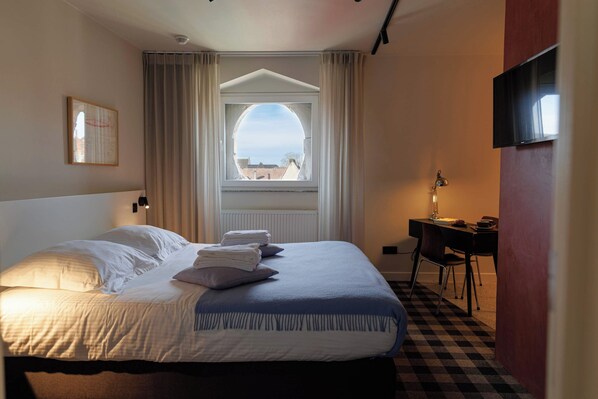 A hotel room with a bed, a desk, a chair, a window with curtains, and a flat-screen TV mounted on the wall.