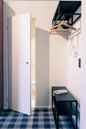 Un couloir avec un sol à carreaux, une porte blanche, une table noire sur laquelle se trouve un magazine, et un cintre en bois sur une étagère noire.
