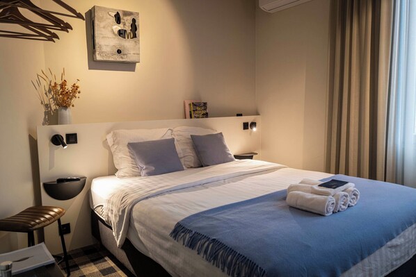 A neatly made bed with a blue blanket and pillows, a bedside table with a lamp, a wall-mounted light, and a small plant on the nightstand.