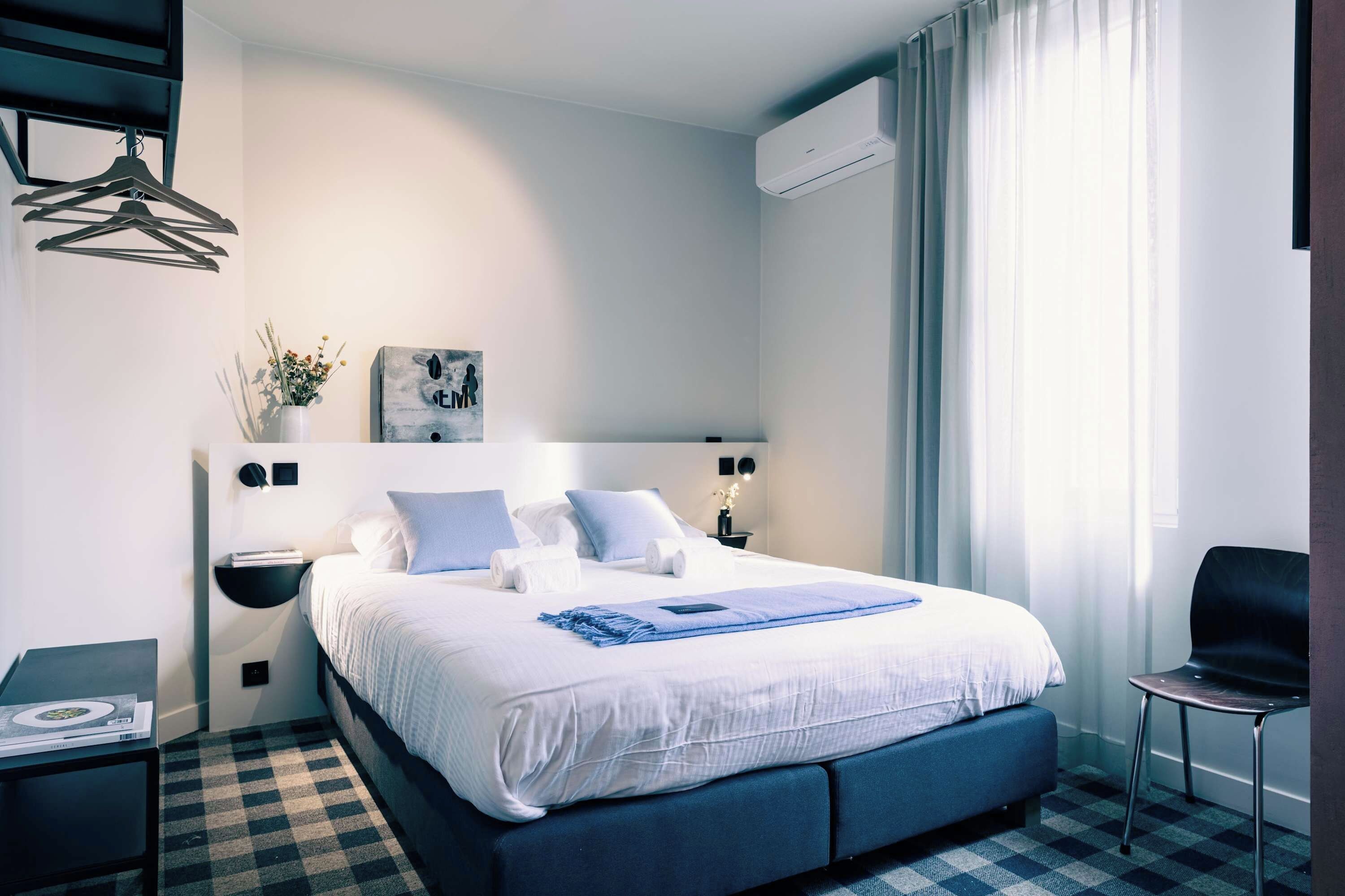 A hotel room with a large bed, a bedside table, a chair, and a wall-mounted air conditioner.