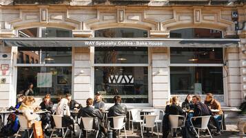 An outdoor cafe with tables and chairs, a sign reading "WAY Specialty Coffee - Bake House," and a building facade with decorative elements.