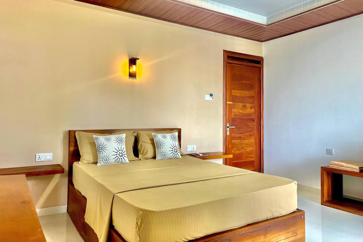 A bedroom with a bed, a wooden headboard, a wall-mounted light, a wooden door, and a small wooden shelf.