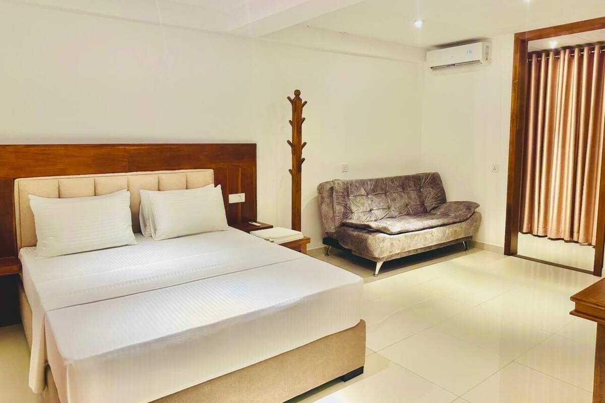 A hotel room with a large bed, a sofa, a wooden coat rack, and an air conditioning unit.