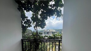 A balcony with a black railing, a wicker chair, and a view of a building and trees in the background. - Easy Stay (Kandy)