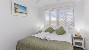 Een slaapkamer met een bed, twee nachtkastjes, een raam met luiken, een lijst met een afbeelding aan de muur, en een groen tapijt met reliëf.