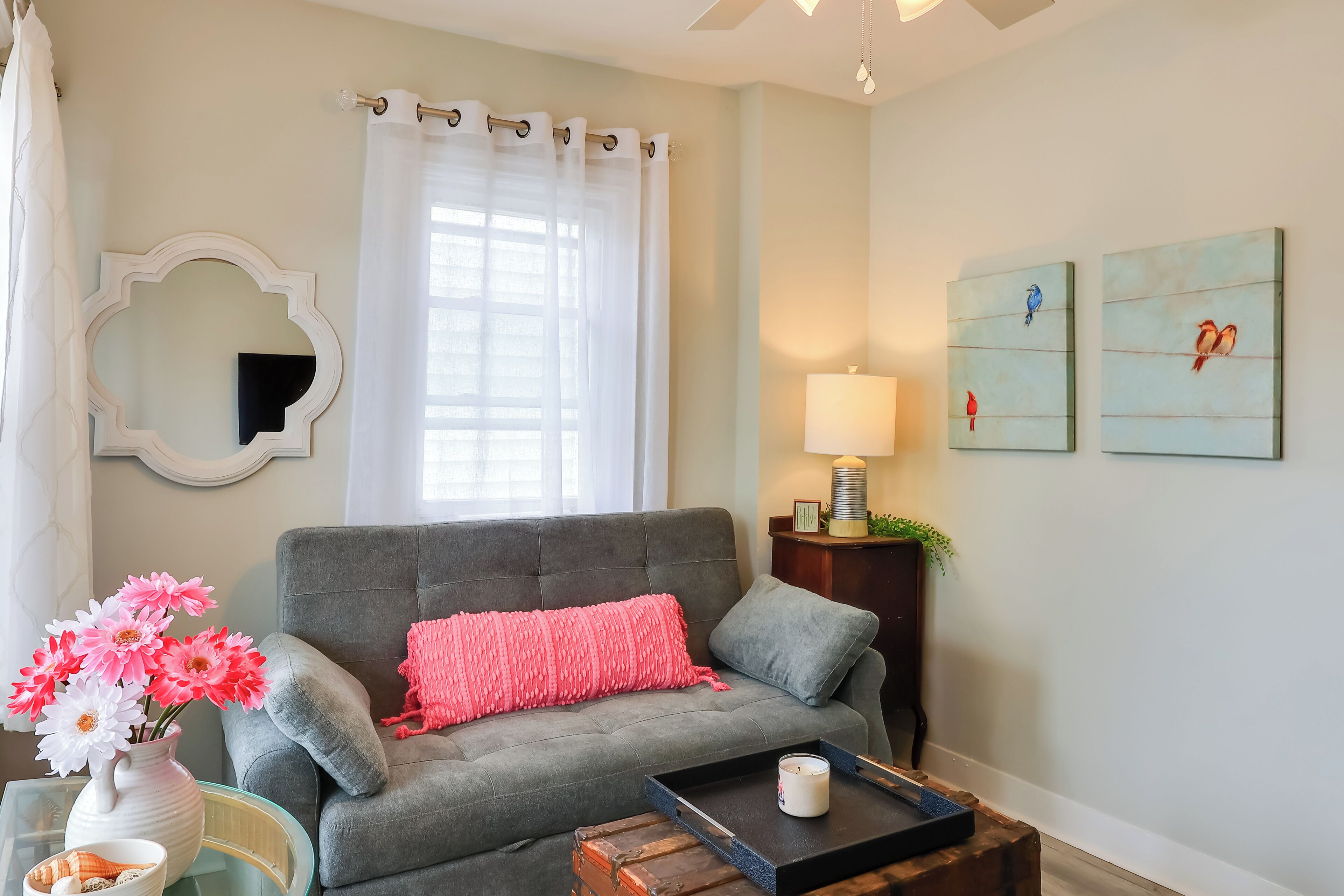 A living room with a grey sofa, a pink throw pillow, a vase with flowers, a lamp, and two wall art pieces.