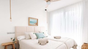 Une chambre avec deux lits, une table de chevet, un ventilateur de plafond, un tableau encadré au mur et une fenêtre avec des rideaux légers.