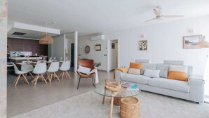 Un salon moderne avec un canapé gris, une table basse en verre et un coin repas comprenant une table et des chaises blanches.