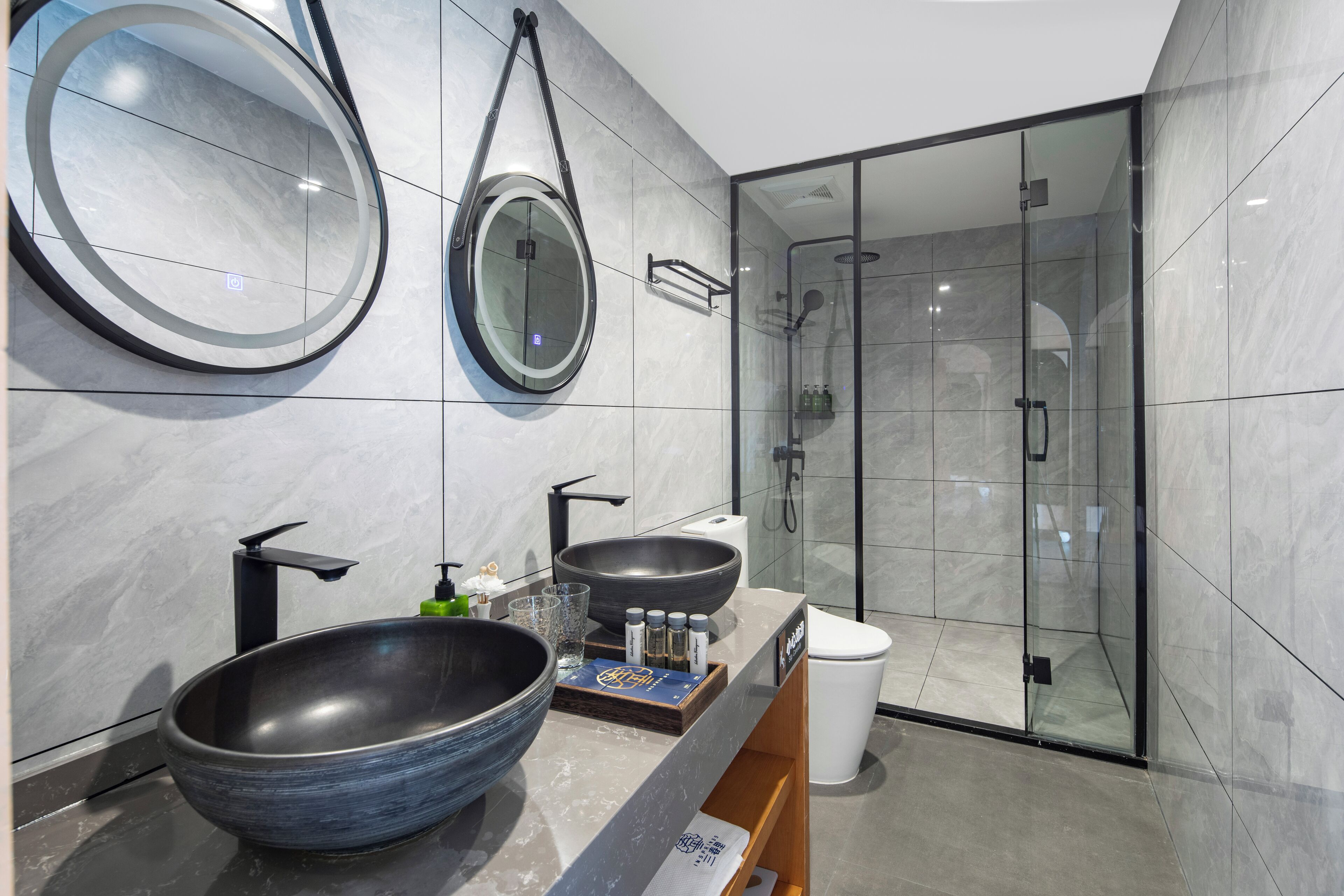 A modern bathroom with two large oval mirrors, a black vessel sink, and a glass-enclosed shower.