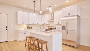 A modern kitchen with white cabinetry, a central island with stools, stainless steel appliances, and pendant lighting. - 2BR/2BA Riverfront Condo w/ Deck (Cashmere)