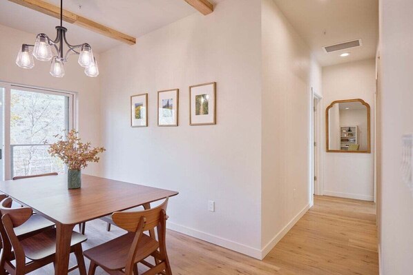 A dining area with a wooden table and chairs, a chandelier, framed pictures on the wall, and a window with a view.