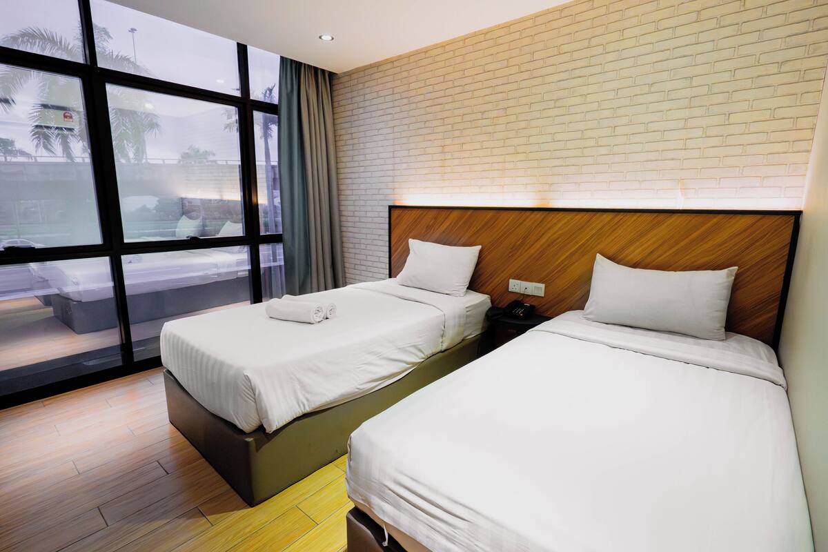 Een hotelkamer met twee bedden, een houten hoofdbord, een raam met uitzicht op palmbomen en een bakstenen muur.