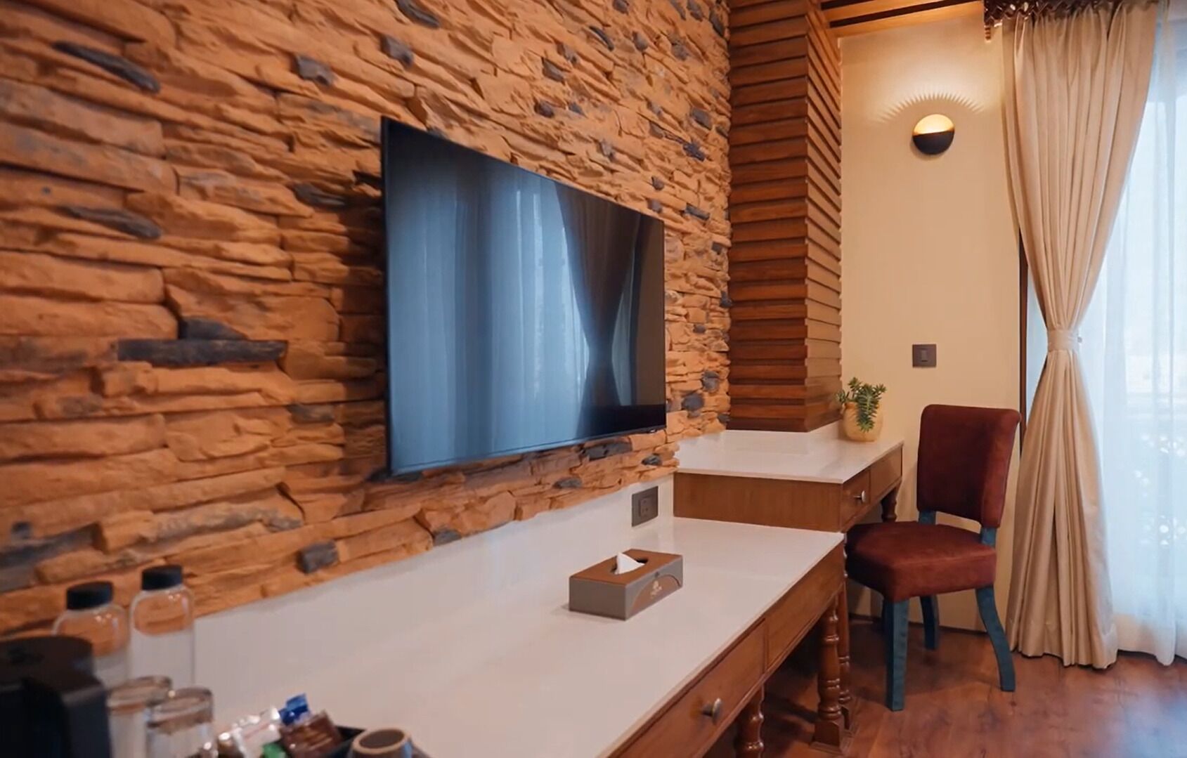 A modern room with a stone wall, a flat-screen TV, a wooden desk, a red chair, and a window with curtains.