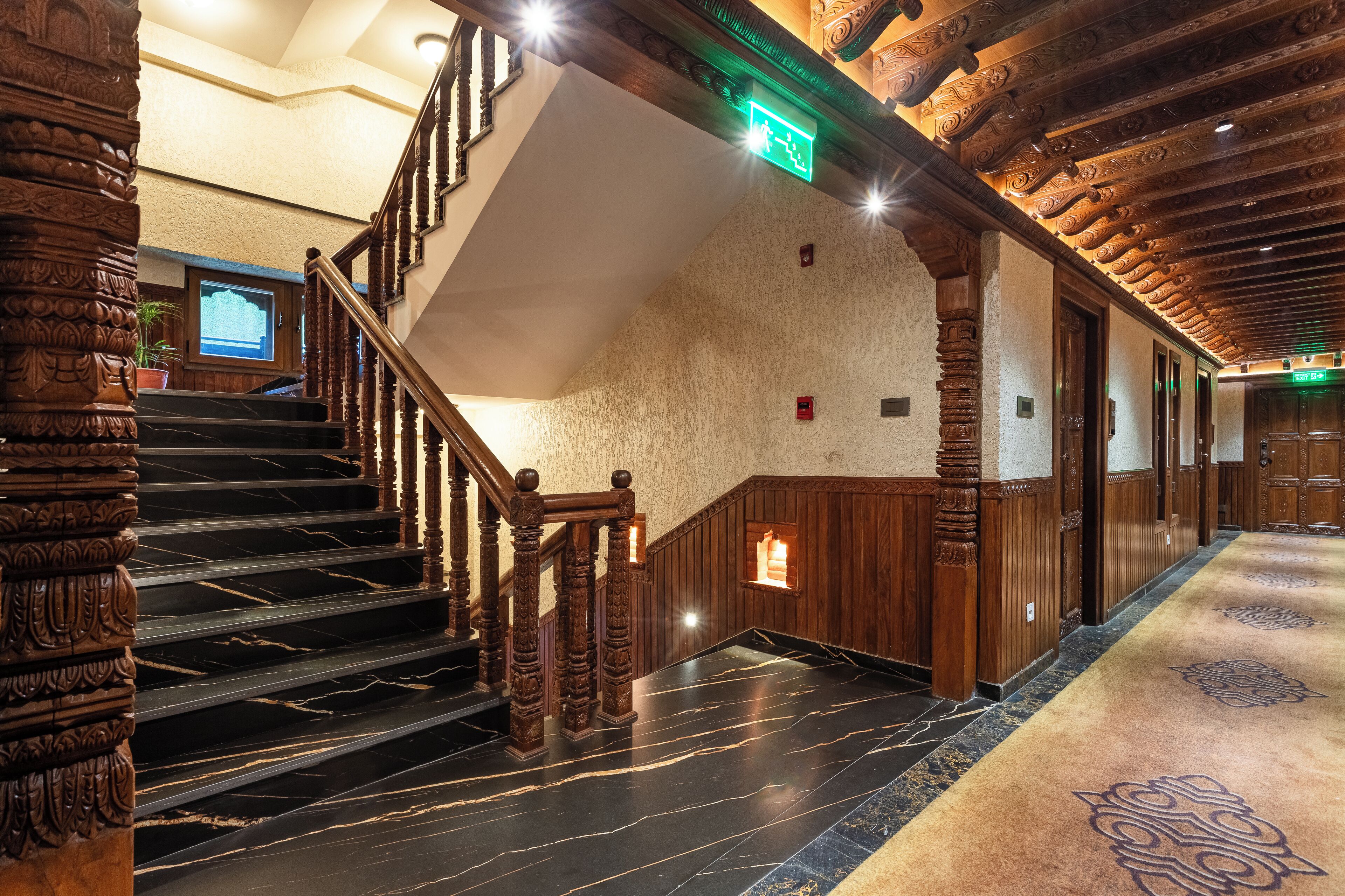 A grand staircase with wooden railings and marble steps, leading to a landing with a green exit sign.