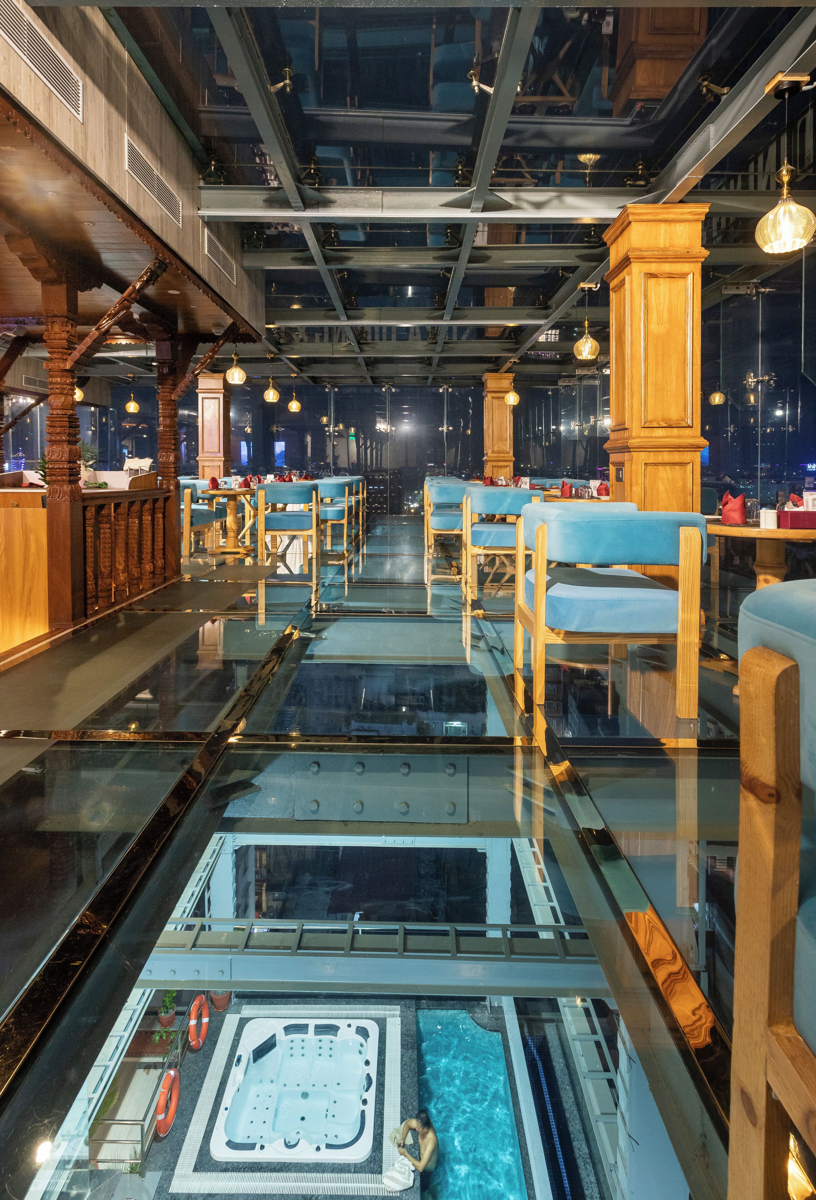 Ein modernes Interieur mit Glasboden, der zu einem Whirlpoolbereich führt, Holzmöbeln und einer hohen Decke mit freiliegenden Balken.