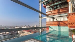 Une piscine à débordement située sur le toit, avec un jacuzzi, entourée de rampes de verre et offrant une vue panoramique sur la ville.