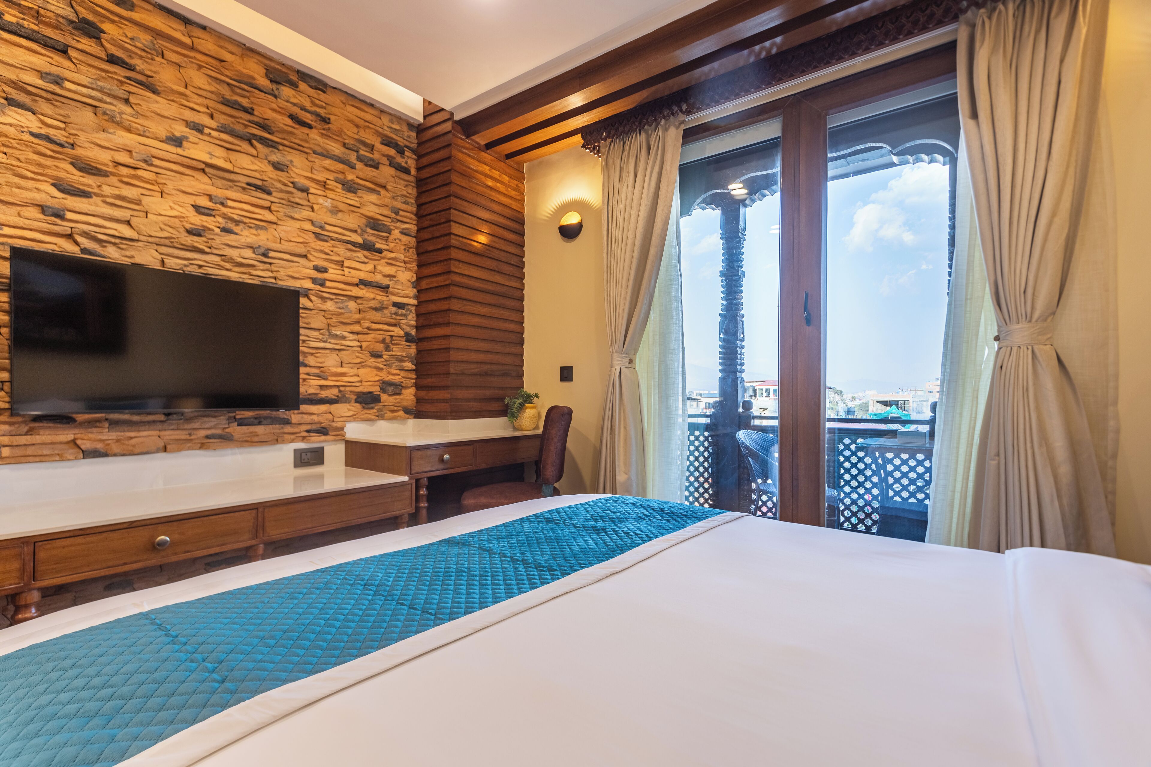 Une chambre d’hôtel avec un mur en pierre, un téléviseur à écran plat, un bureau, un lit recouvert d’une courtepointe bleue et un balcon avec des rideaux.