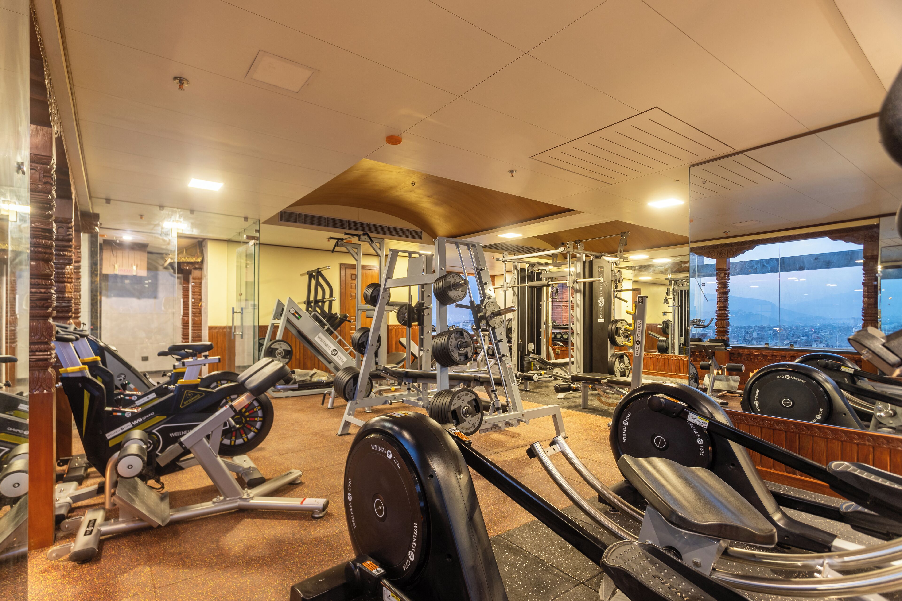 Ein Fitnessstudio mit verschiedenen Trainingsgeräten, darunter Laufbänder und Kraftgeräte, einem großen Spiegel, der die Geräte und einen Blick auf eine Stadtlandschaft reflektiert.