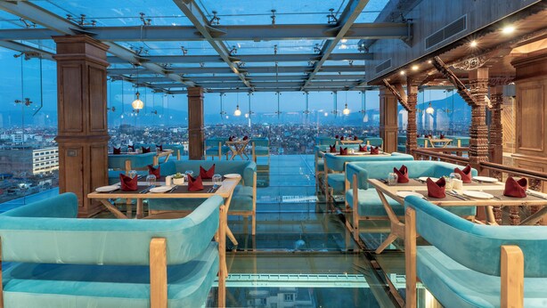 Un restaurant situé sur le toit d’un immeuble, doté d’un plancher en verre, de sièges rembourrés bleus et offrant une vue sur la ville.