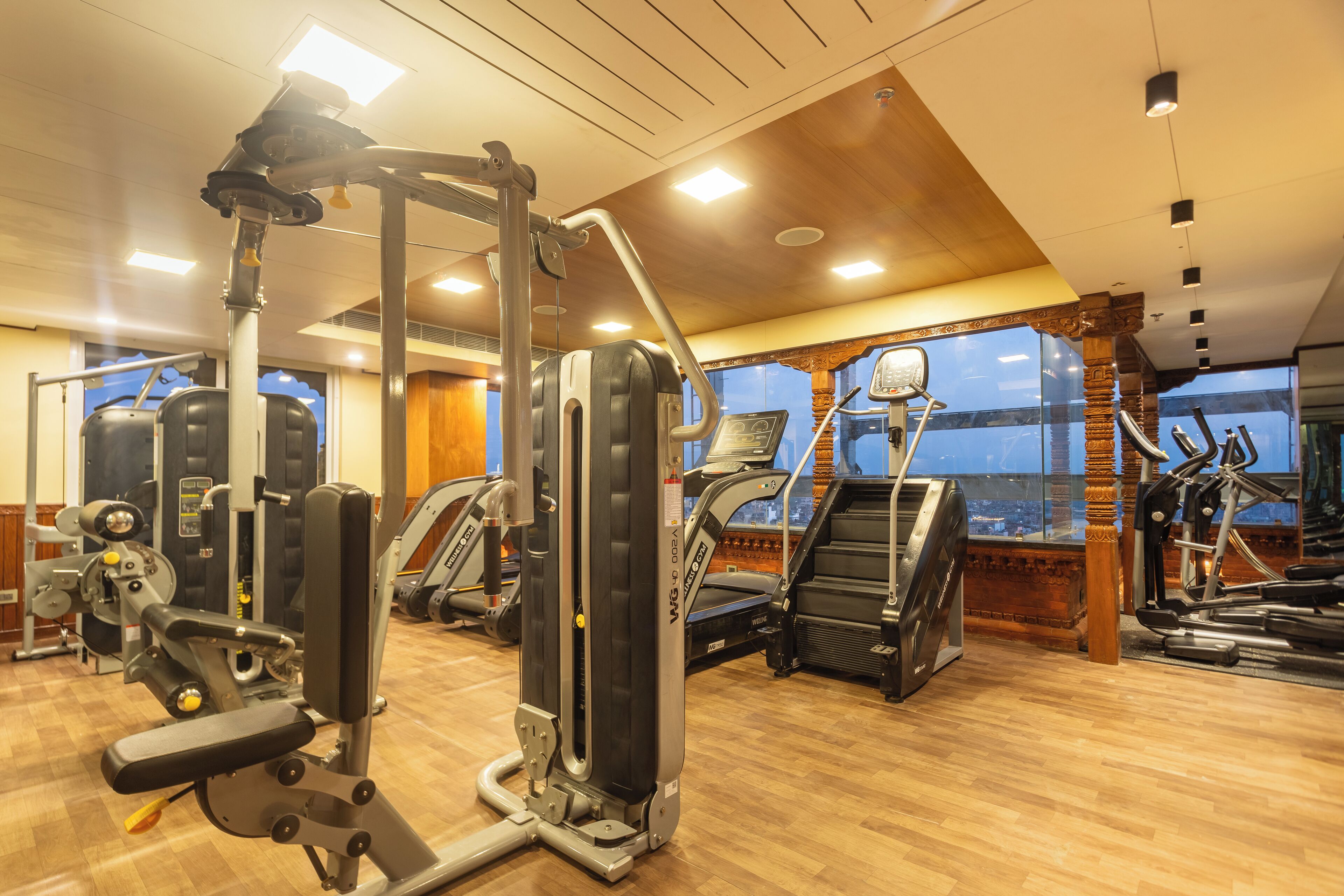 A gym with various exercise machines, wooden flooring, and large windows offering a view of the city.