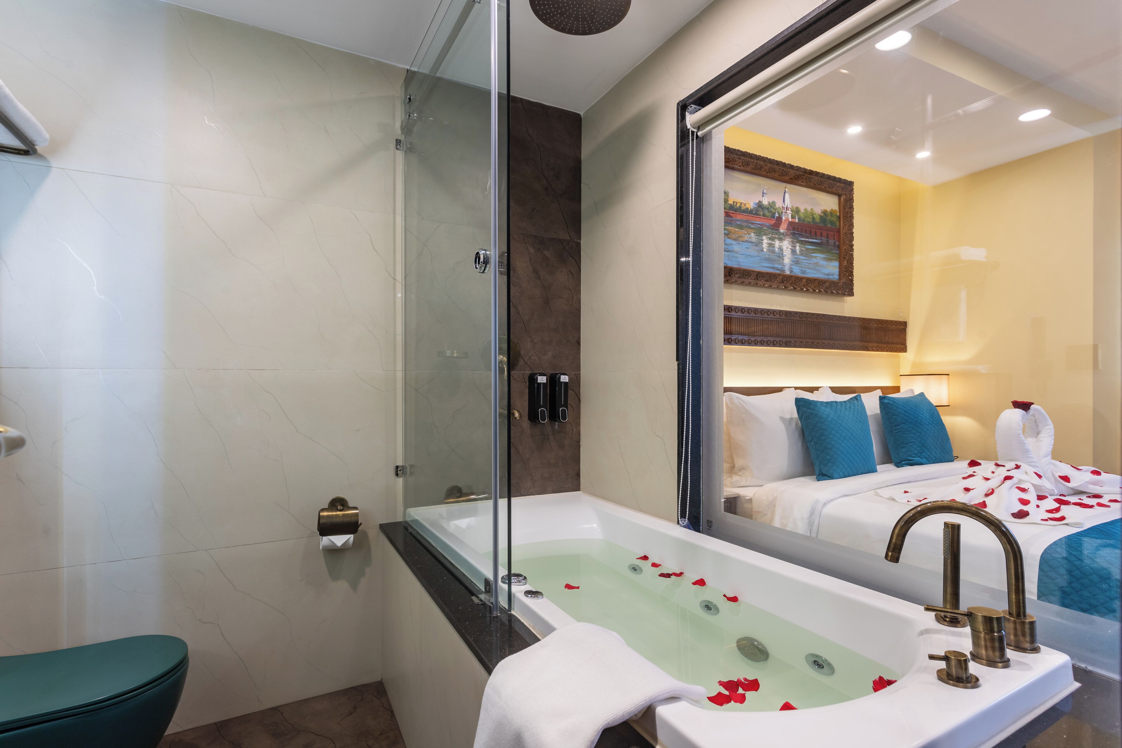 A modern bathroom with a glass-enclosed shower, a large bathtub with rose petals, and a bed with blue pillows and a white blanket.