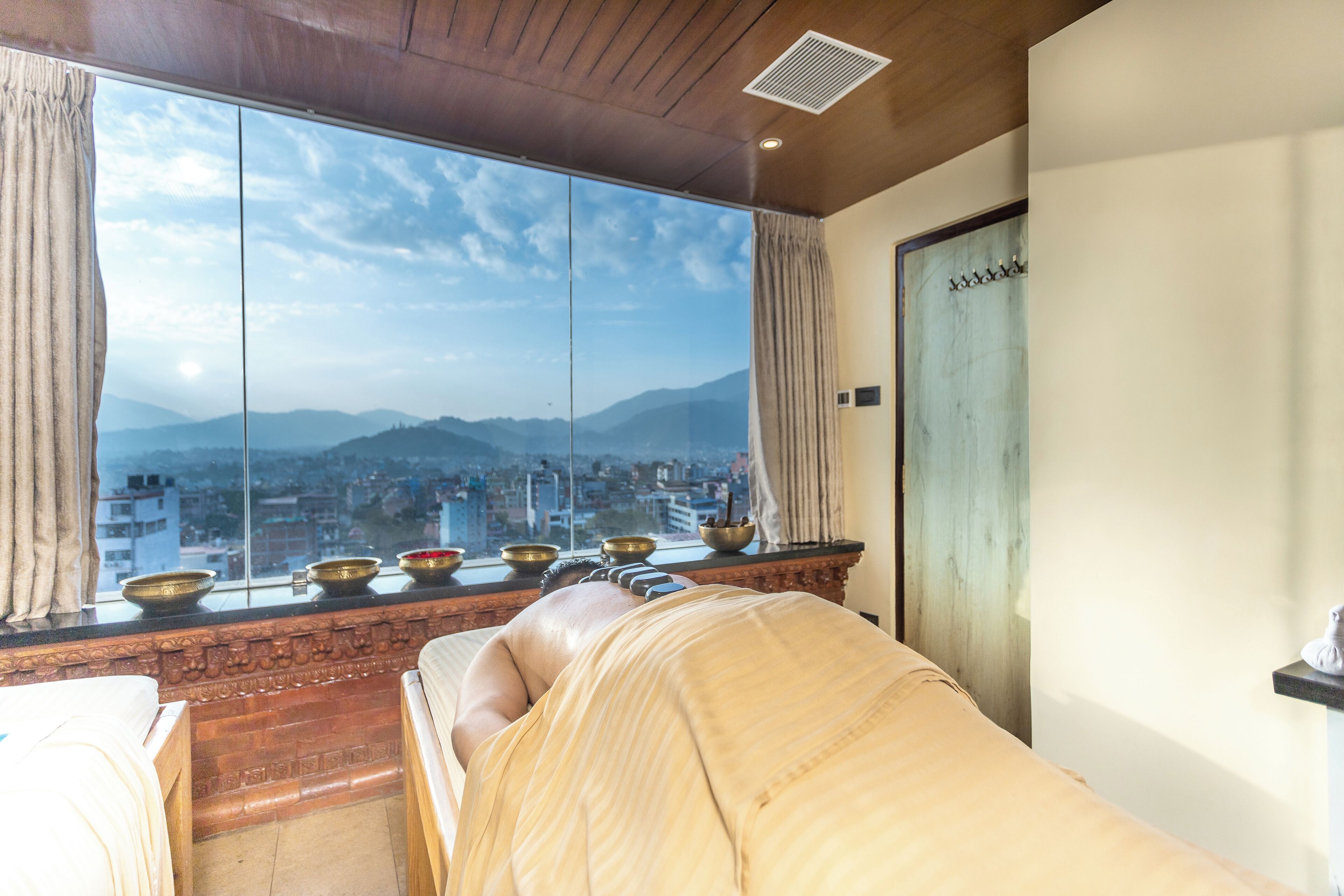 A room with a large window offering a city view, a massage table, and a shelf with bowls.