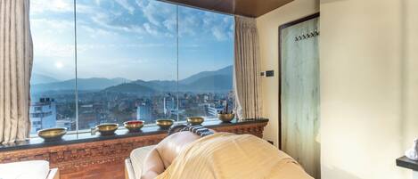 A room with a large window offering a city view, a massage table, and a shelf with bowls.