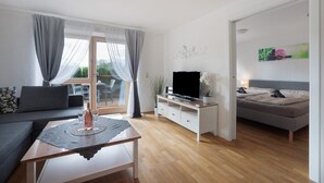 A modern living room with a sofa, a TV on a stand, a dining table with a placemat, and a bed with a headboard.