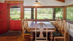 Un comedor con una mesa y sillas de madera, ventanas amplias con vista a un paisaje verde y una puerta roja.